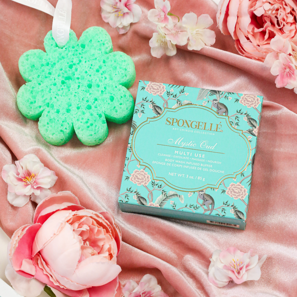 Green flower-shaped body buffer sponge beside Mystic Oud multi-use box on fabric