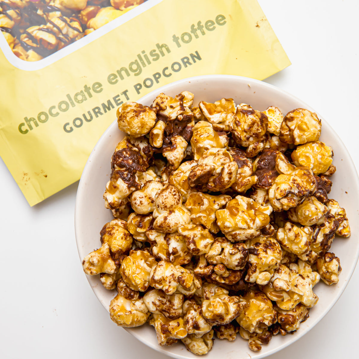 Bowl of chocolate toffee popcorn with mixed golden and dark coated pieces, beside yellow bag
