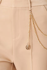 Camel woven pants close-up: gold-tone drape chain and round charm at pocket