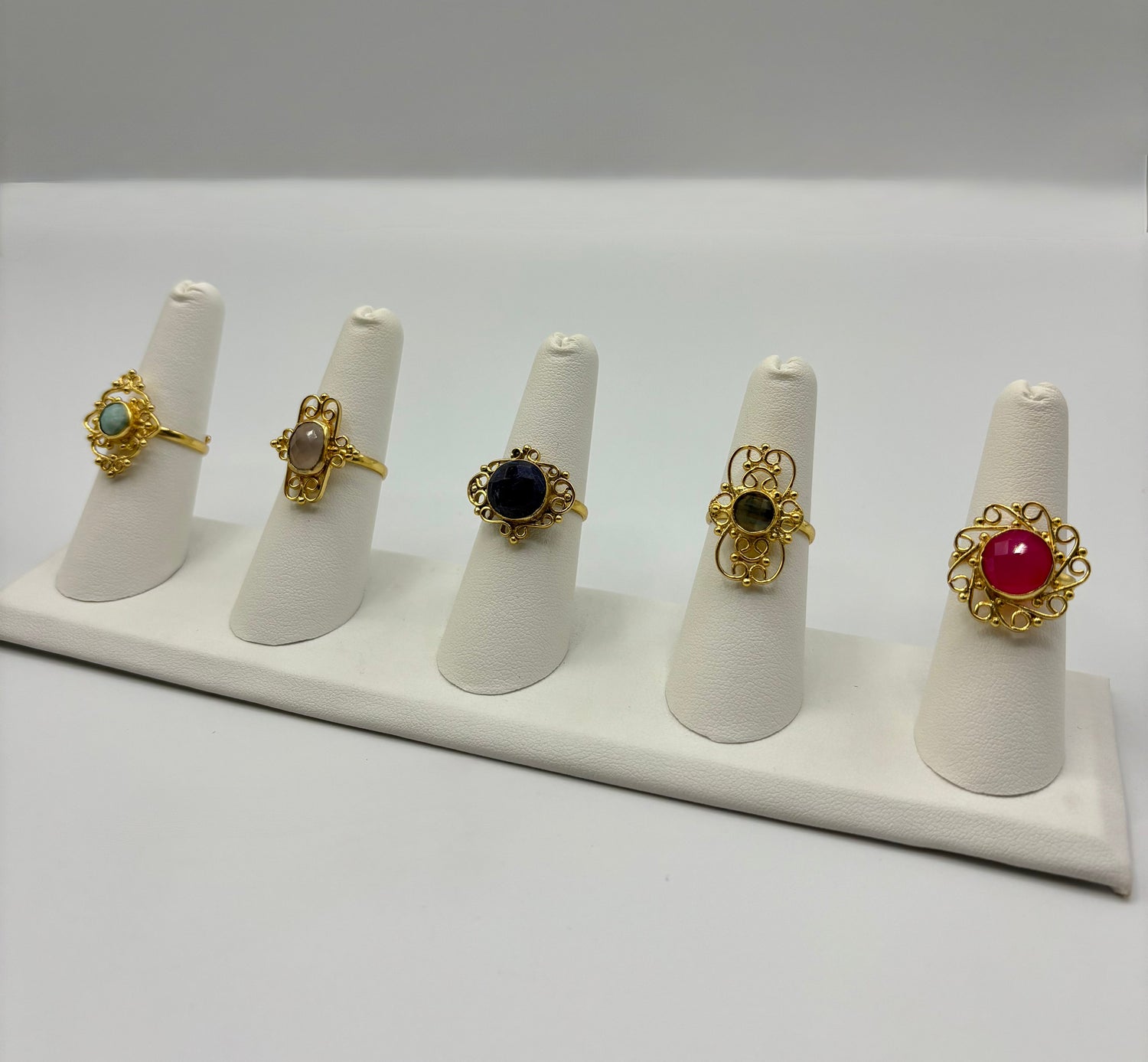 Gold-tone gemstone rings on white stands: blue, black, green, and red round stones