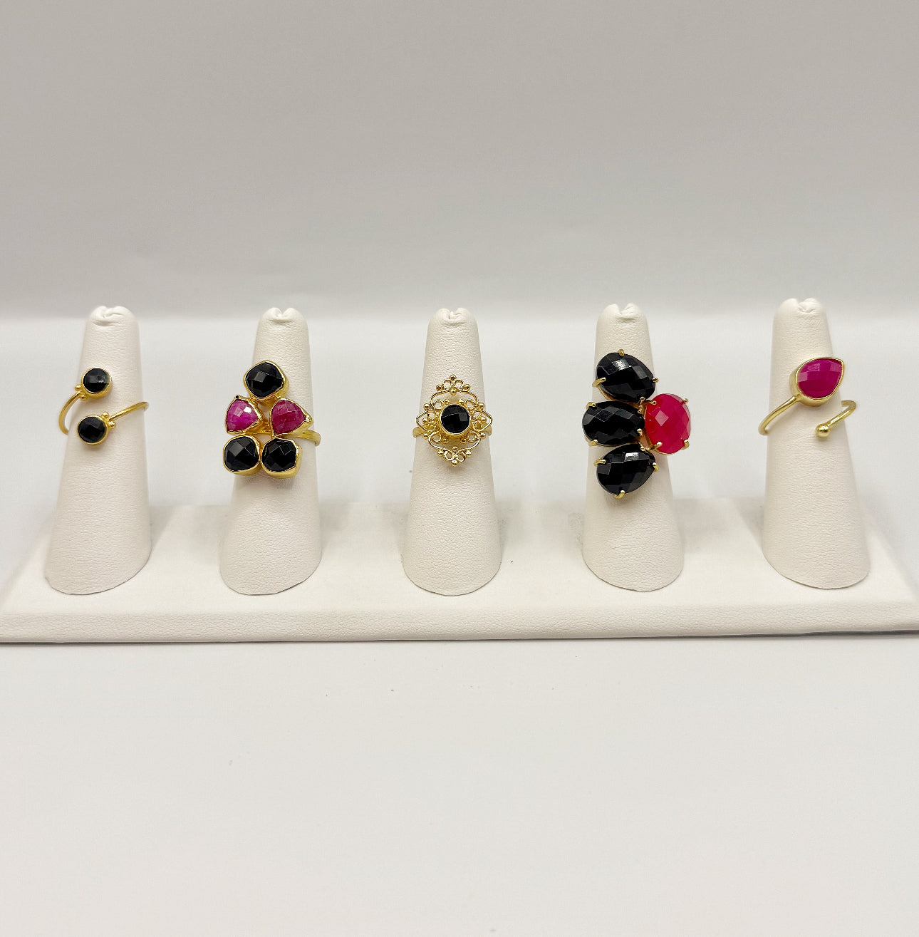Five gold-tone gemstone rings with black and hot-pink stones on white stand