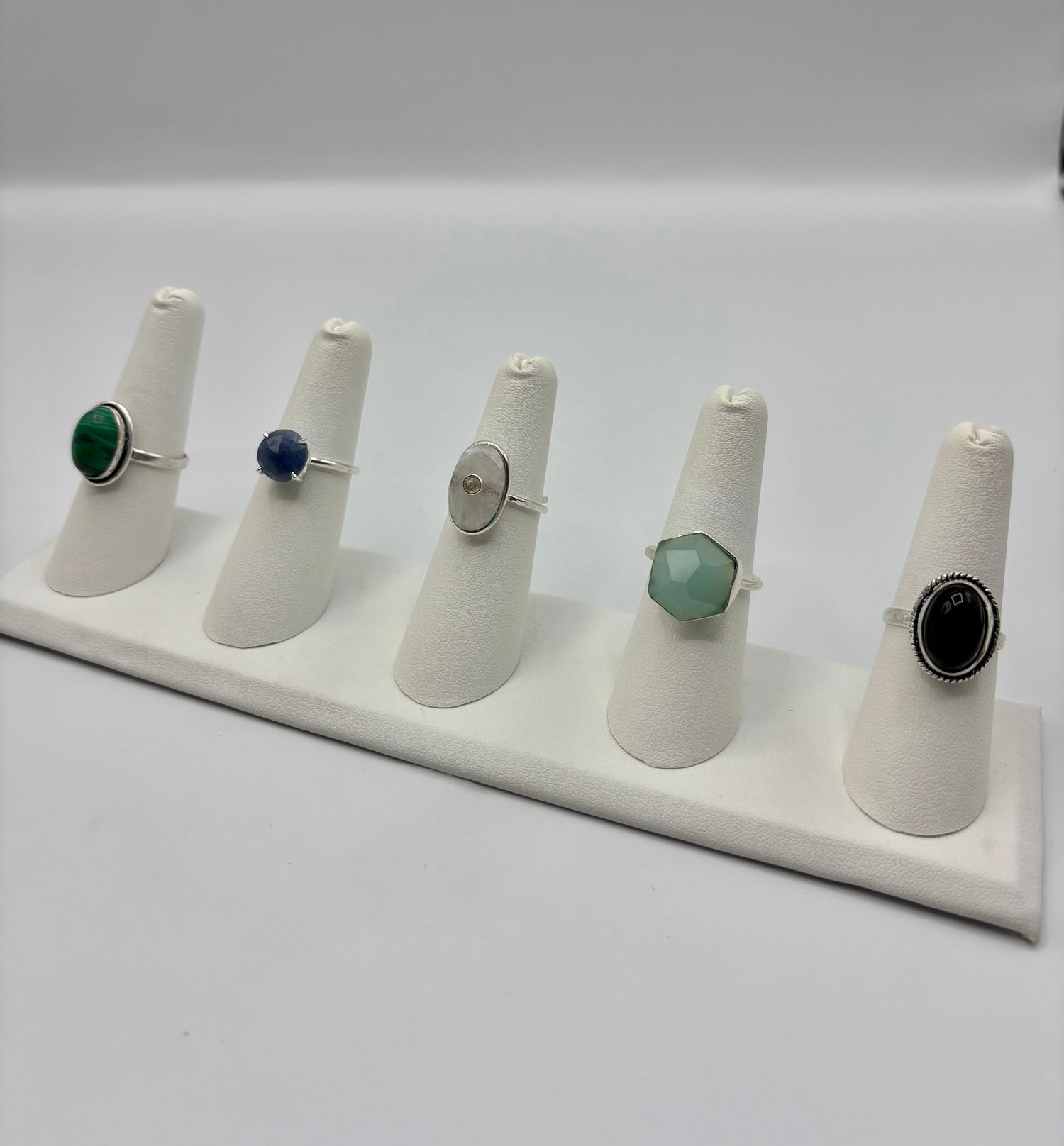 Five gemstone rings displayed on white ring holders, including green and blue stones