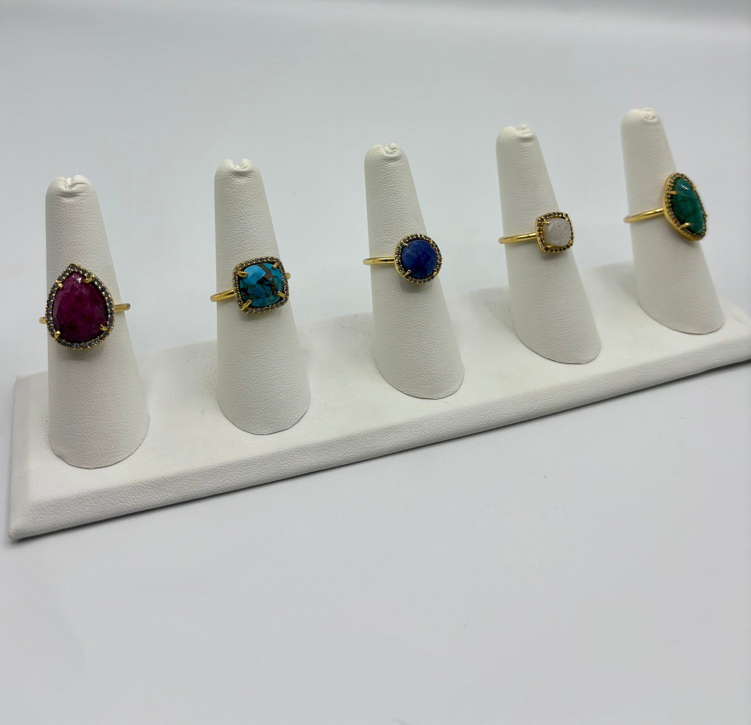 Gold-tone gemstone rings on white display stands with blue, teal, and purple stones