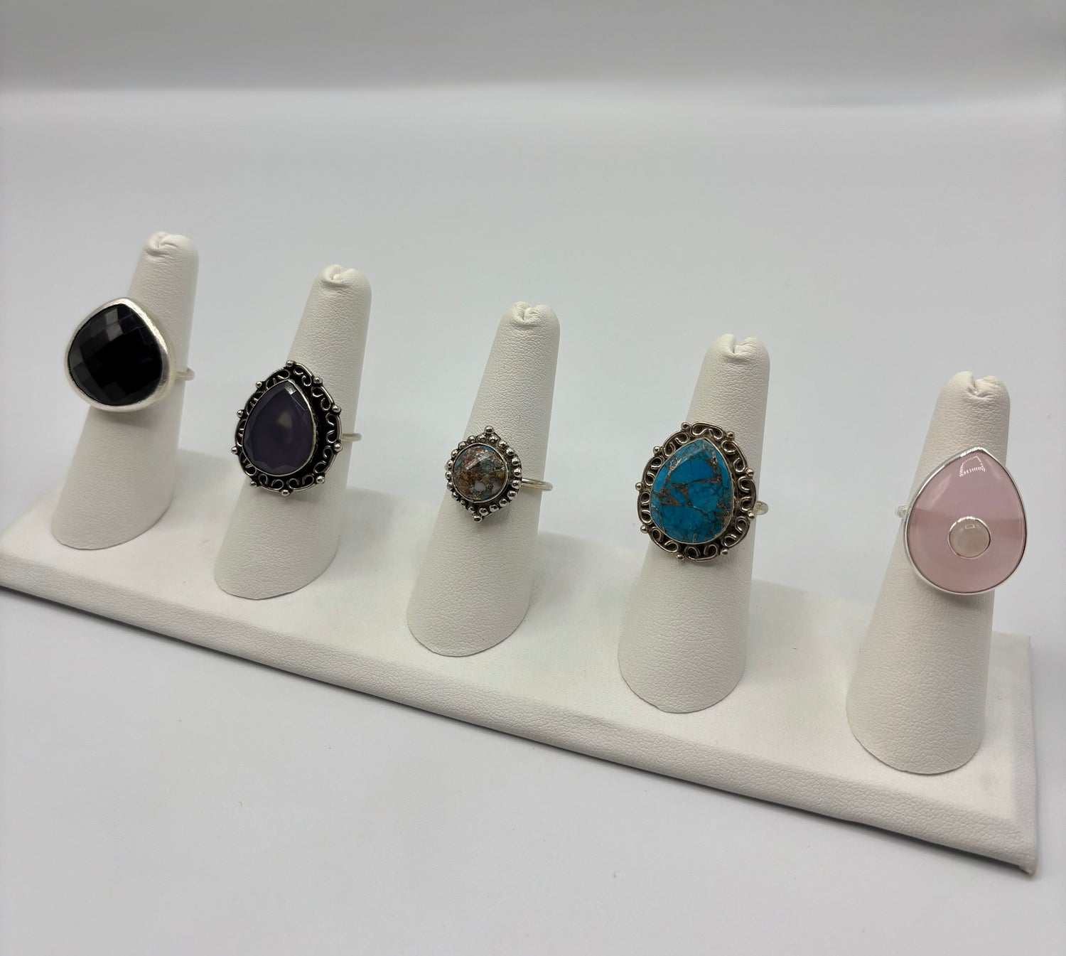Five rings displayed on stands: black, dark purple, silver, turquoise, and rose stones