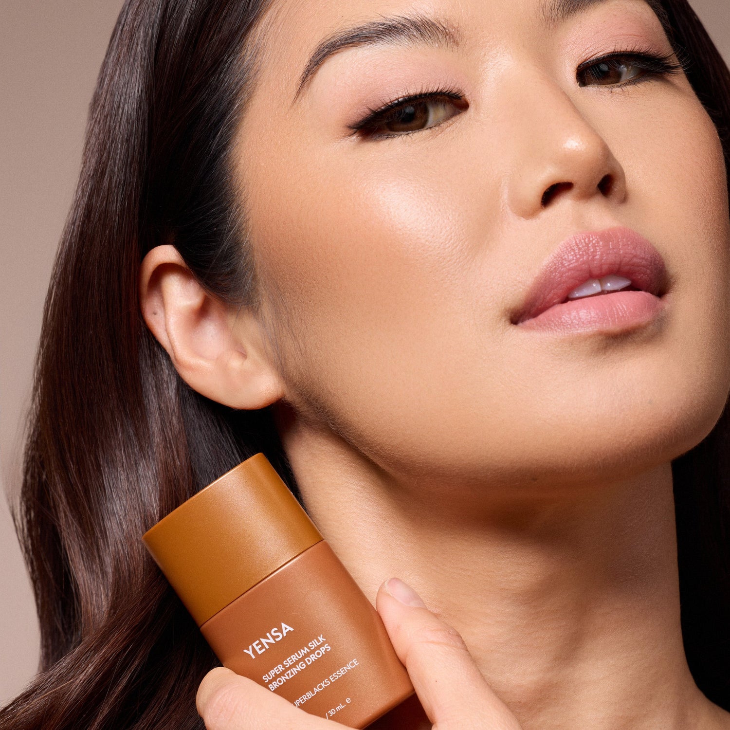 Bronzing serum silk drops bottle held near a woman’s cheek